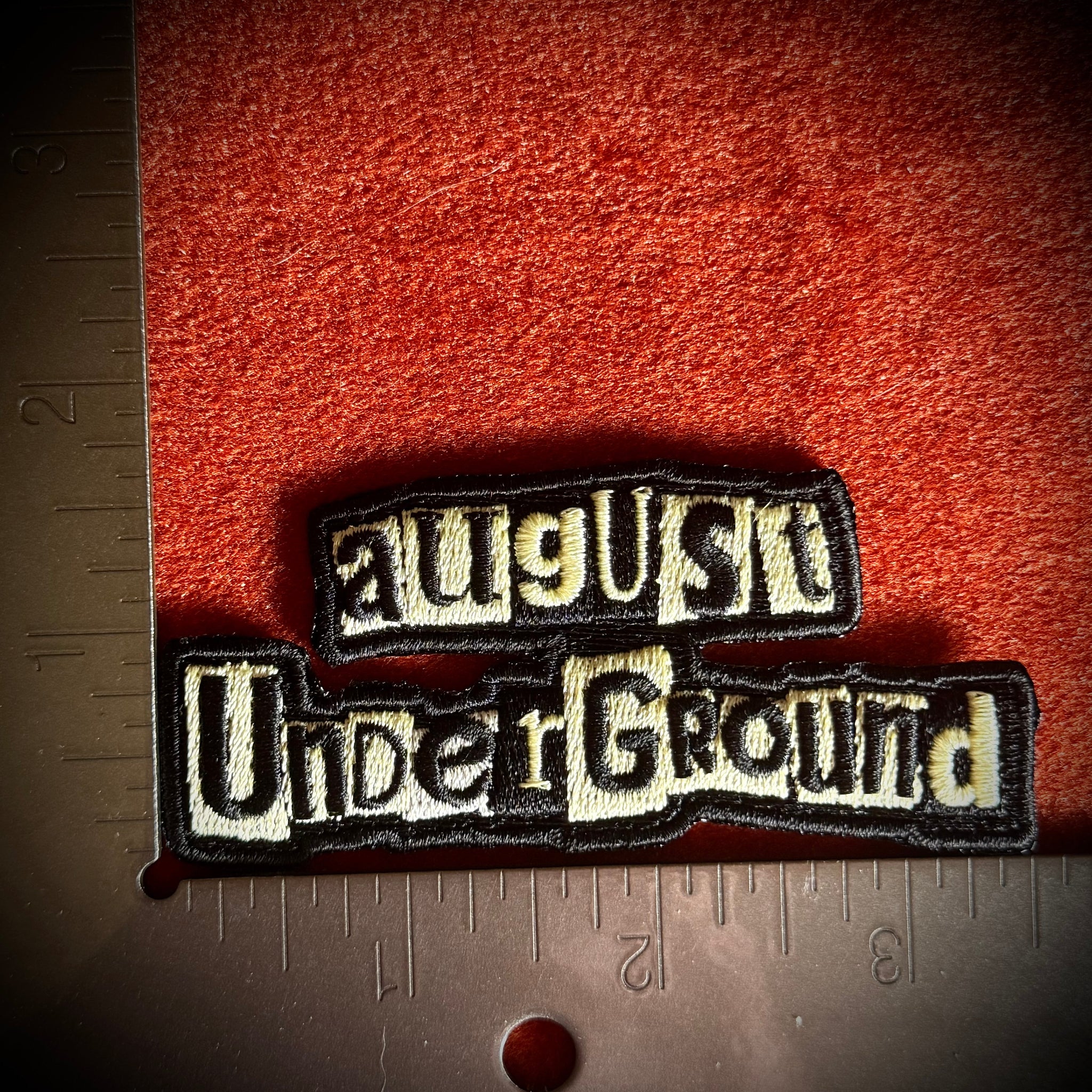 An embroidered patch reading 'August Underground' in cream lettering with a black border next to a metal ruler for scale.