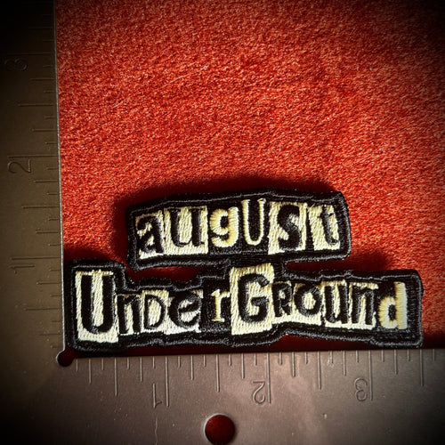 An embroidered patch reading 'August Underground' in cream lettering with a black border next to a metal ruler for scale.