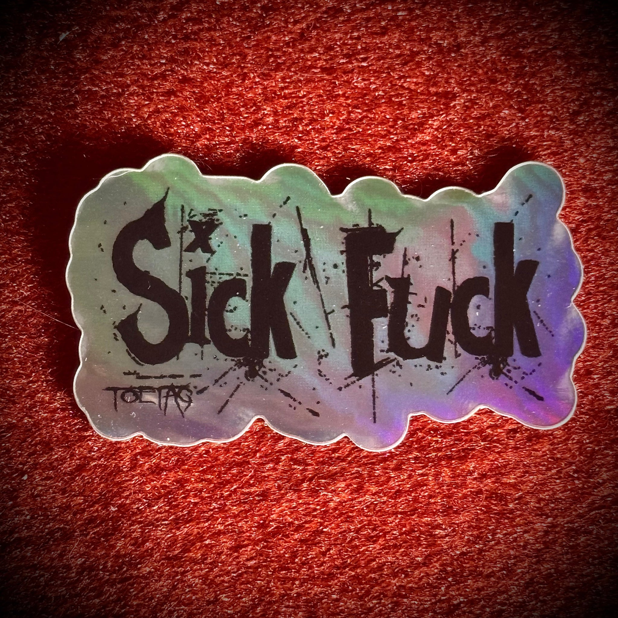 A die-cut sticker with the words 'Sick Fuck' in black distressed lettering over an holographic background.